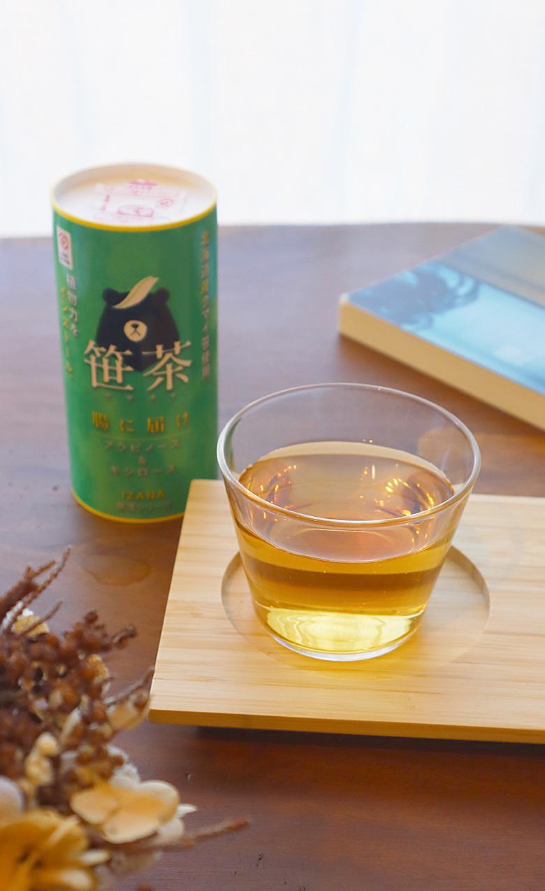 白いカーテンを背景に木目のテーブルがあり、笹茶のボトルと笹茶が注がれたガラスのコップがお盆に置かれている。暖かな日差しが差し込み、全体的に清潔感と親しみやすさが感じられる。