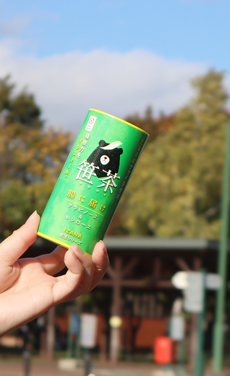 青い空と緑の自然を背景に、笹茶のボトルを持つ人の手が見える。柔らかな自然光が差し込み、明るい雰囲気が漂う。